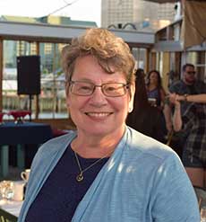 Woman wearing a blue sweater and glasses, standing outside and smiling.