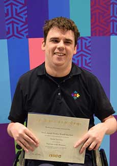 A young man in a wheelchair holds a certificate, smiling. Certificate text is not legible.