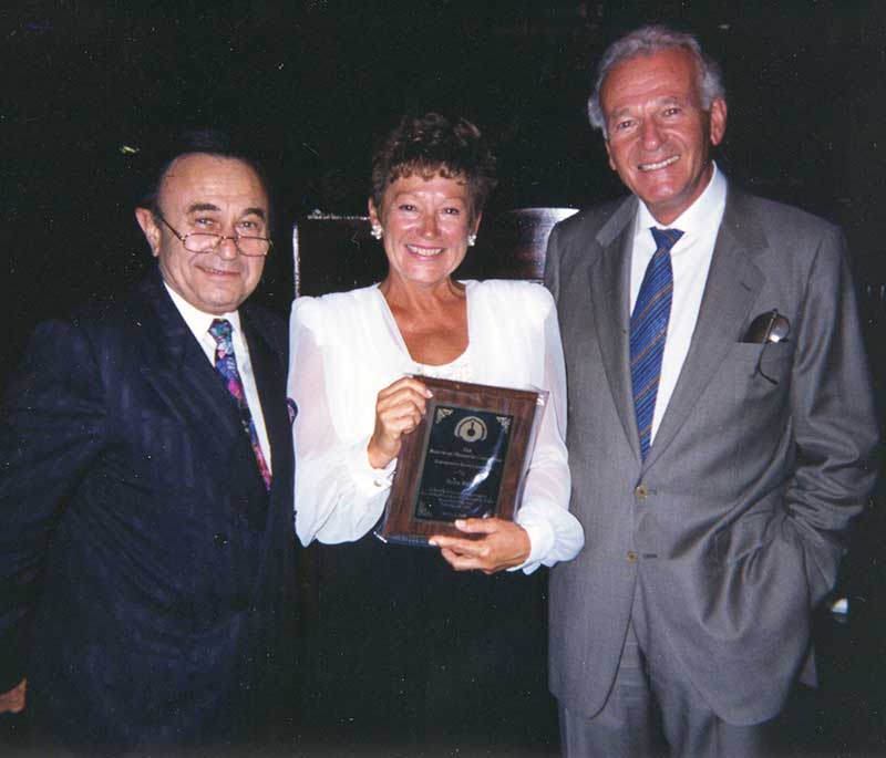 A smiling young woman holds an award while standing with two young men.