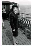 Woman in dark coat standing in front of bench on deck of ship.