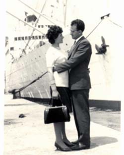 Man and woman hugging each other on pier with ship in background.