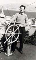 Man holding on to ship's wheel, land and sea in background.