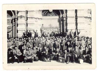 Large group of men seated and standing on steps of concrete building.