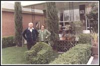 Couple standing on lawn with hedges and green grass.