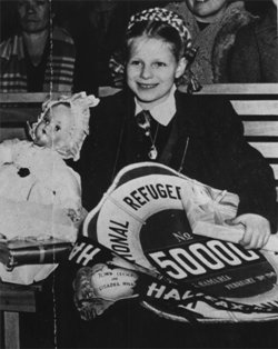 Ausma as a child, sitting next to a doll and holding a sign saying 50 000th Refugee.