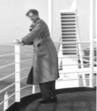 Bernardus in a trench coat leaning up against railings of ship.