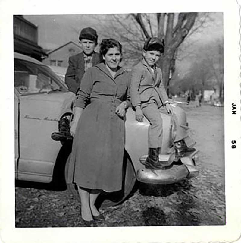 A woman leans against a car with two boys are sitting on the hood.