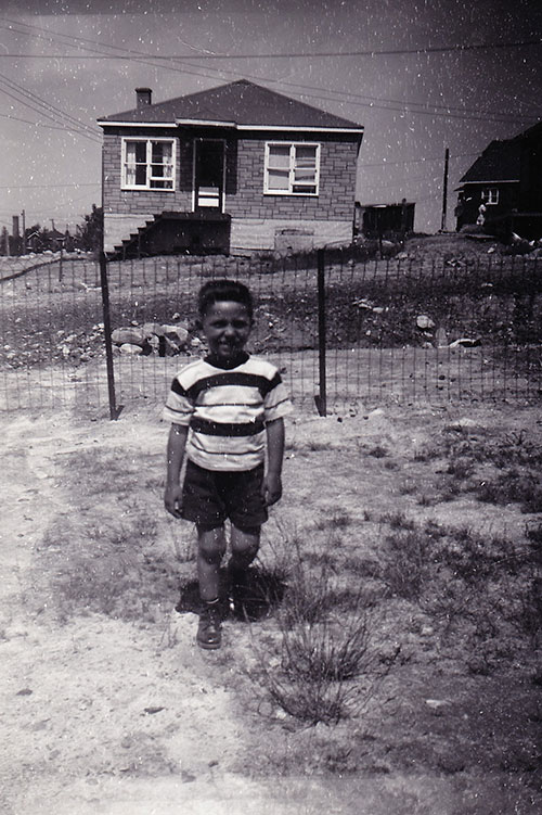 A small boy wearing shorts & t-shirt standing outside of the fence for photo.