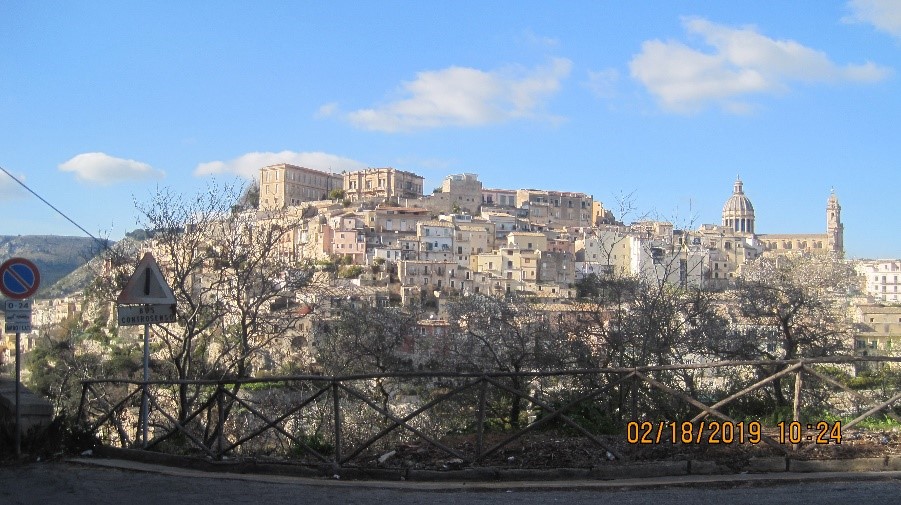 The town of Ibla from a distance, with a barrier and trees in front.