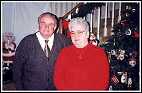 Coloured photo of older man and woman in front of Christmas tree.