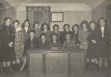 Several young women seated and standing behind a desk.