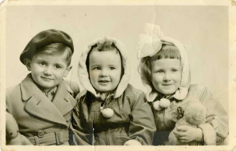 Three small children sit in a row, all wearing very different winter hats.