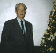 Older man in suit and tie standing next to Christmas tree.