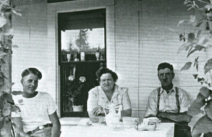 Young Cornelius and older couple sitting at table on porch of house.