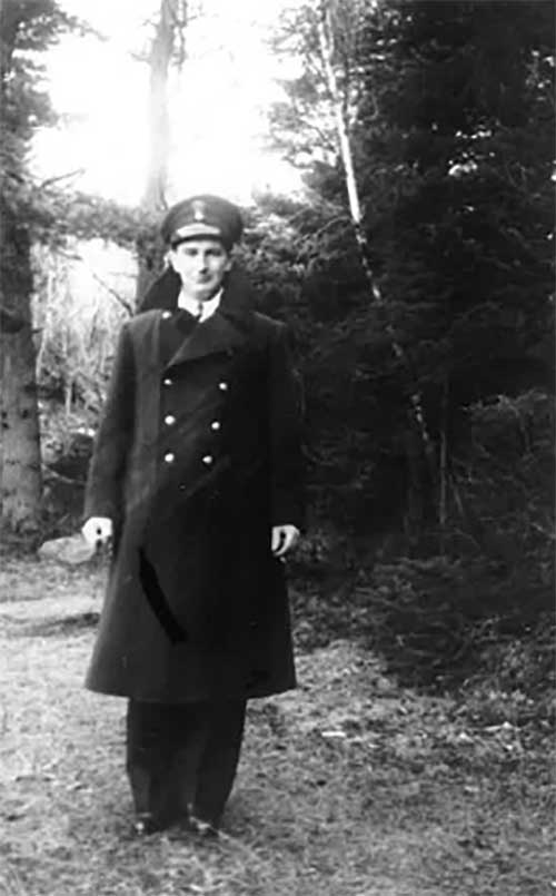 A man in a uniform is wearing a long black coat and a cap, and is standing in front of tree. 