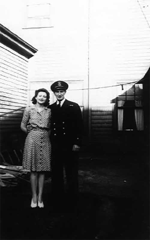 A young couple are standing in front of a house, she wears a nice dress and he is in uniform.