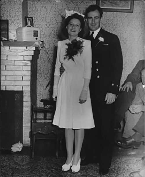 A young couple stand next to a fireplace in wedding attire.
