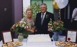 Pompilio and Rosa standing behind table laden with cake, flowers and balloons.