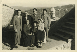 Old outside photo of two women seated with baby, four men standing behind.