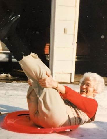 An elderly white woman outside on a circular sled, laughing and holding her legs in the air.