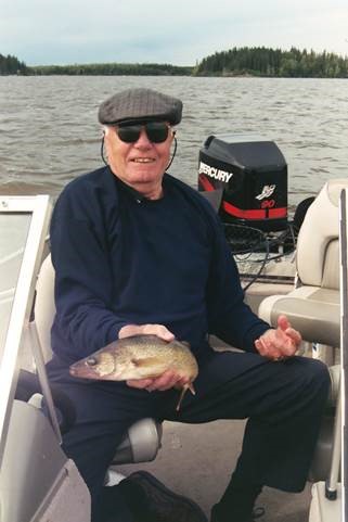 An older white man on a boat on a lake, holding a fish and smiling at the camera.
