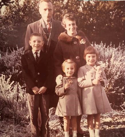 A sepia-toned photo of a white family including a man, woman, and three children, standing outside and dressed nicely.