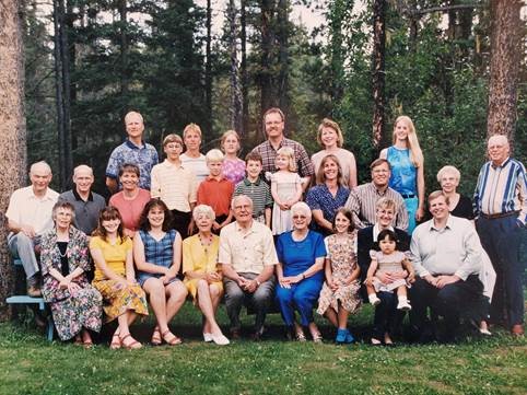 A large, posed group photo of a white family, taken outdoors in warmer weather with a forest behind them.