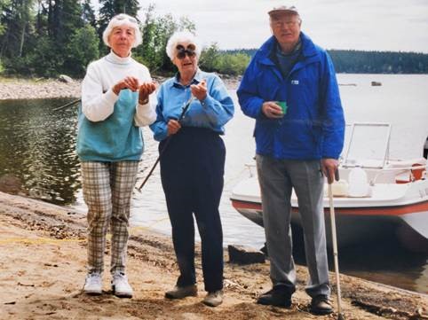 Three older white people, two woman and one man, standing by a lake with sticks and a boat behind them.