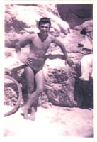 Young Eddy in swimming trunks, leaning against rocks on beach.