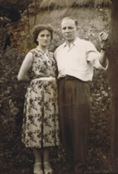 A young couple are standing in front of bushes as they have their photo taken.