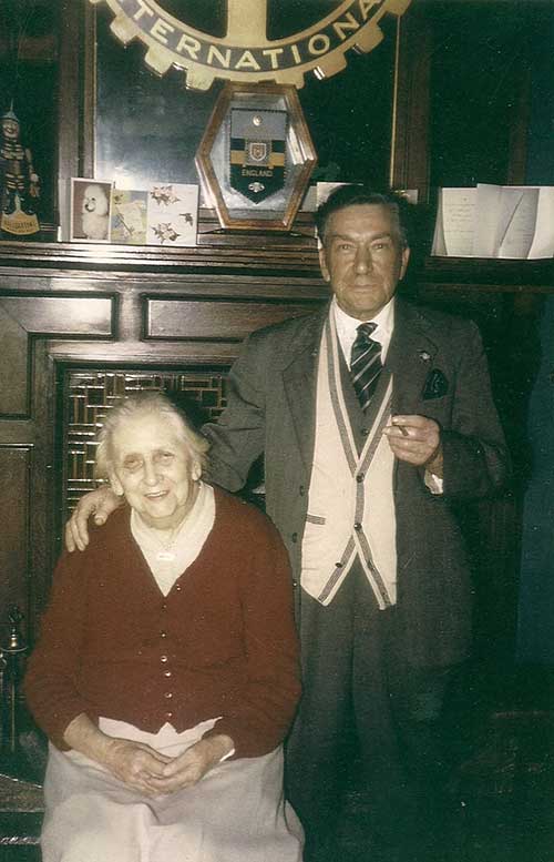 An elderly woman is sitting on a chair, a man stands next to her in front of a fireplace.