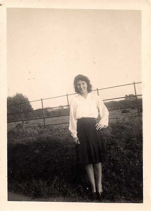 A young woman stands in front of a hedge. 