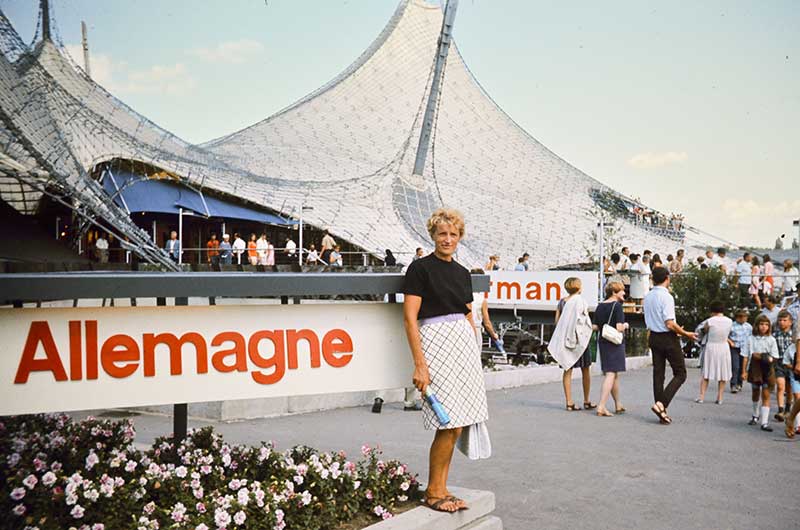 A young woman stands in front of the Munich Olympics Stadium, the word Allemagne is spelled out in big letters next to her.