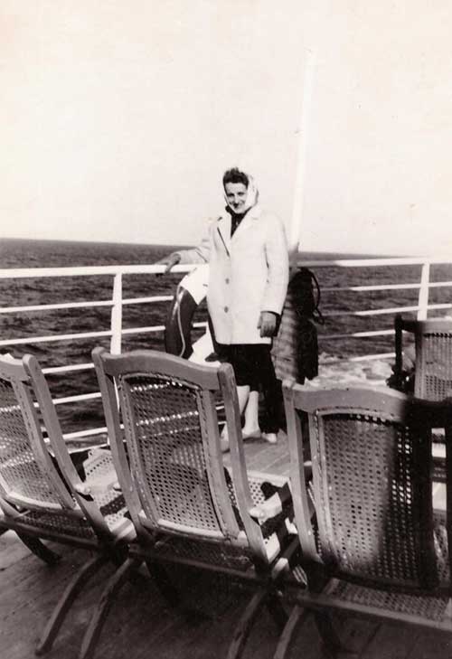 A young woman in a winter coat and kerchief leans against the railing on a ship’s deck.