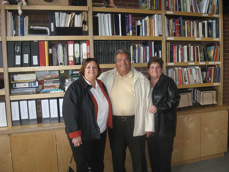 A young man with two women are standing next to a big book shelf.