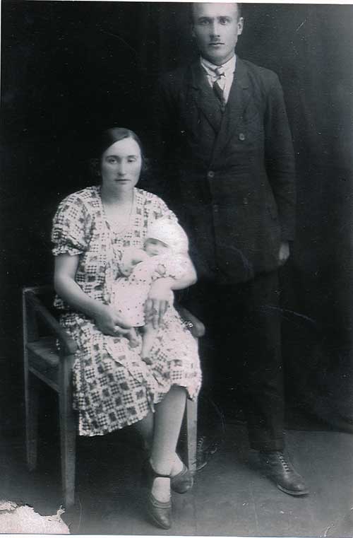 Archival image of a woman seated with a baby in her arms, a man in black stands next to her.
