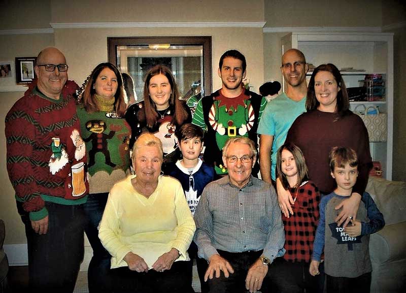 Young family members wearing Christmas sweaters circle an elderly couple who are seated.