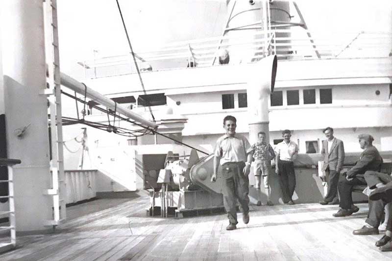 Young men can be seen on the deck of a ship, one of them is walking toward the camera.