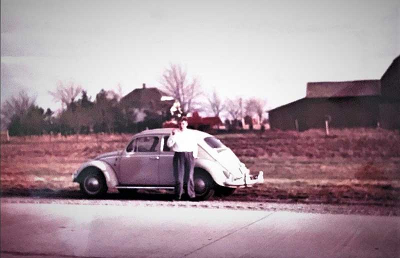 A young man leans against a white Volkswagen Beetle and looks across the road at the camera.