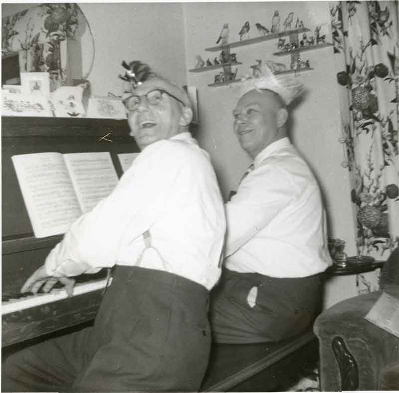 Two gentleman sit at a piano, laughing and singing.
