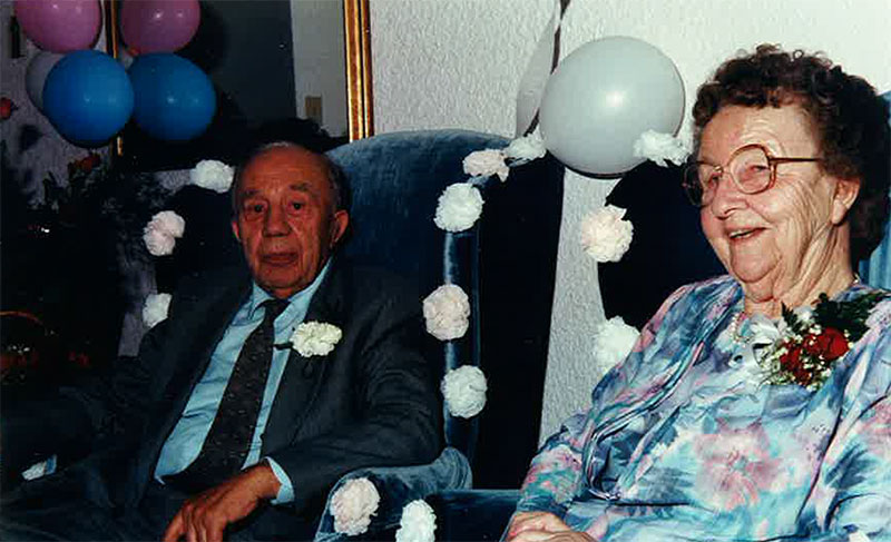 A young couple is sitting on wingback chairs that are decorated with white flowers and balloons.