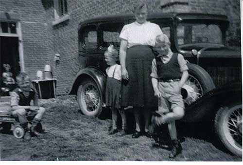 Two children and a woman are  standing in front of a car.