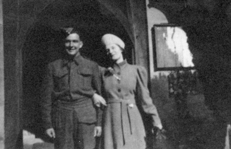 A young man stands in military uniform, with a woman next to him holding his arm.