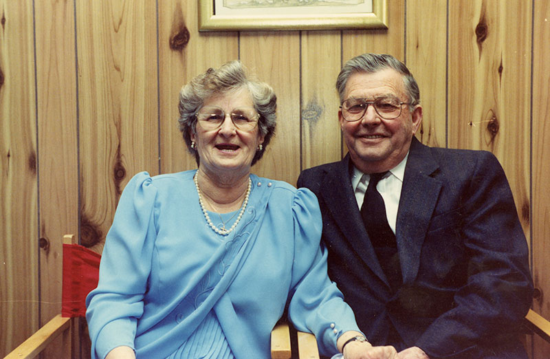 Woman in a light blue dress and wearing pearls sits next to a man in a navy blue suit.