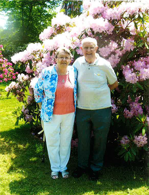 An elderly couple are standing in front of a beautiful flowering bush as they pose for a photo.