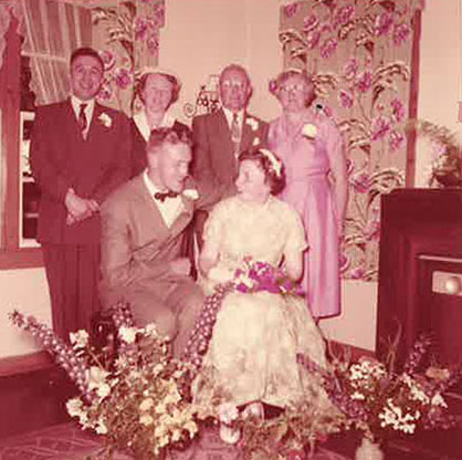 A newly married couple are seated and four guests stand behind them.