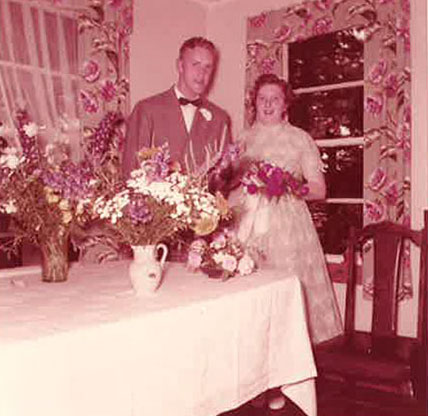 A newly married couple stand behind a table laden with food and drink.