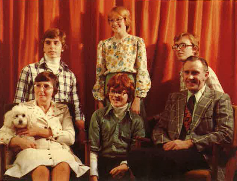 A family of six are in front of an orange curtain as they pose for a photo, one woman is holding a dog.