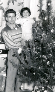 Older photo of man with small child, standing next to Christmas tree.