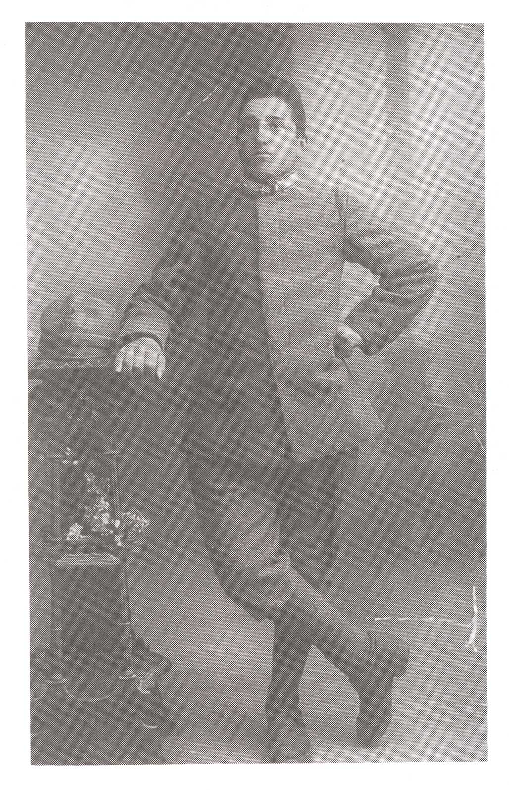 Very old image of a young boy standing with his legs crossed and his hand rests on a shelf.